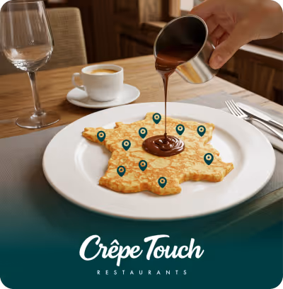 Une crêpe en forme de carte de France avec plusieurs icônes de localisation, du chocolat chaud est versé dessus à côté d'une tasse de café et d'un verre vide sur une table en bois.